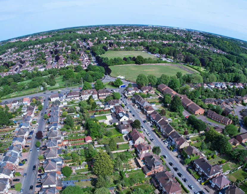 A quiet residential street during daytime with multi-storey brick houses lining the left side, each with small front gardens bordered by neatly trimmed bushes and plants, including a small purple-flow